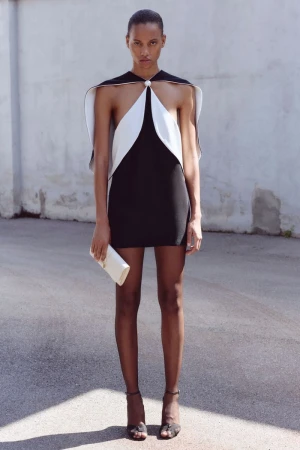 Model wearing a black mini dress with a white draped capelet collar and holding a white clutch outdoors.