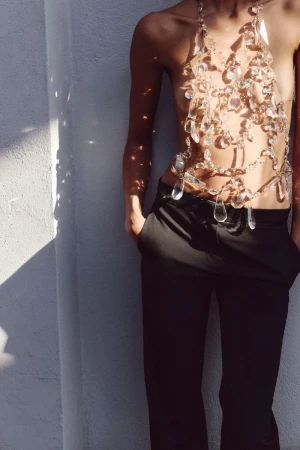 Model wearing an intricate crystal halter body piece and black tailored trousers against a sunlit white wall.