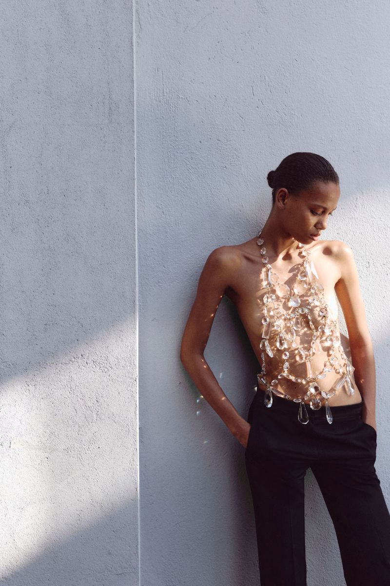 Model standing against a light textured wall wearing black tailored trousers and an intricate clear crystal body harness.