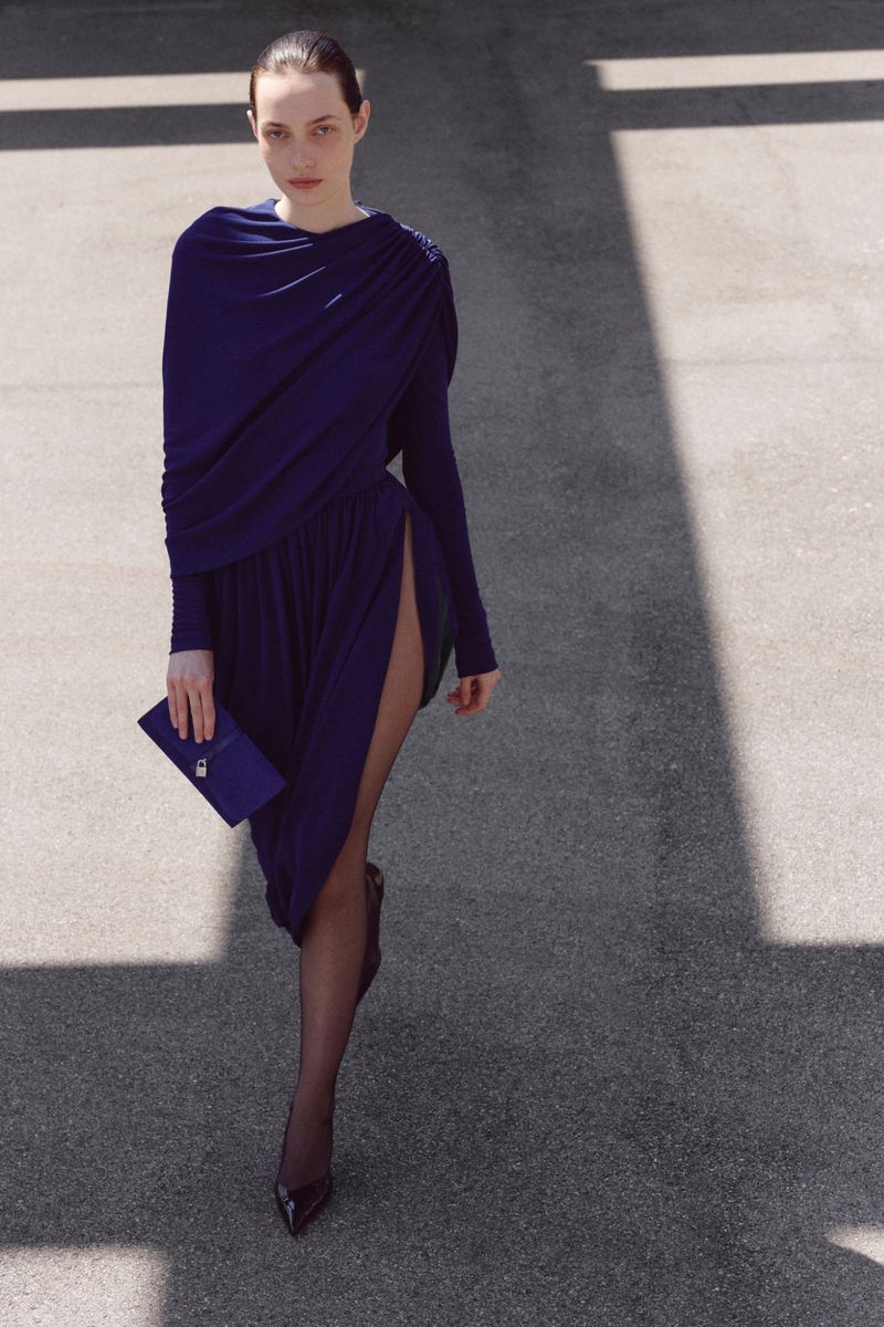 Model walking forward in a draped navy blue long-sleeve dress with a high side slit, sheer tights, and a blue clutch bag.