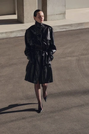 Model walking forward on asphalt wearing an oversized high-collar black shiny coat, sheer tights, and pointed black heels.