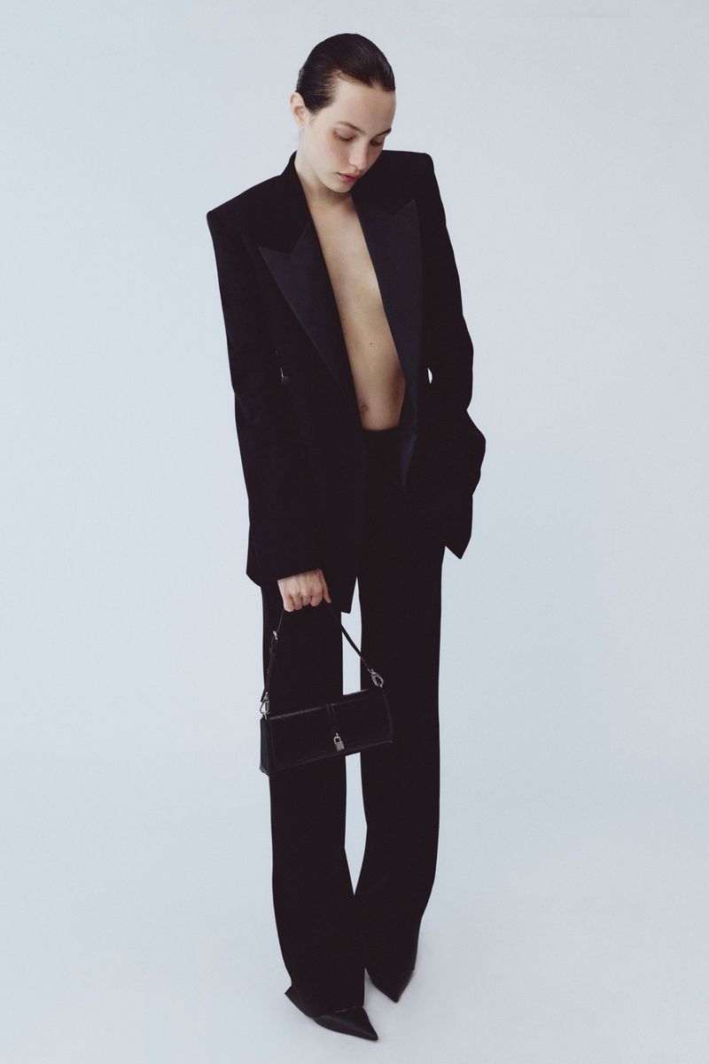 Model wearing a black tailored blazer and trousers holding a small black handbag against a light background