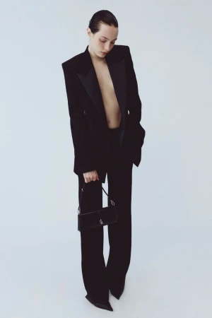 Model wearing a black tailored blazer and trousers holding a small black handbag against a light background