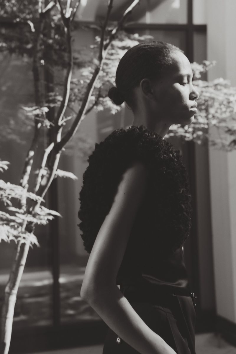 Model in profile wearing a dark textured sleeveless top beside an indoor tree in a black and white portrait.