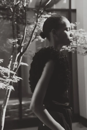 Model in profile wearing a dark textured sleeveless top beside an indoor tree in a black and white portrait.