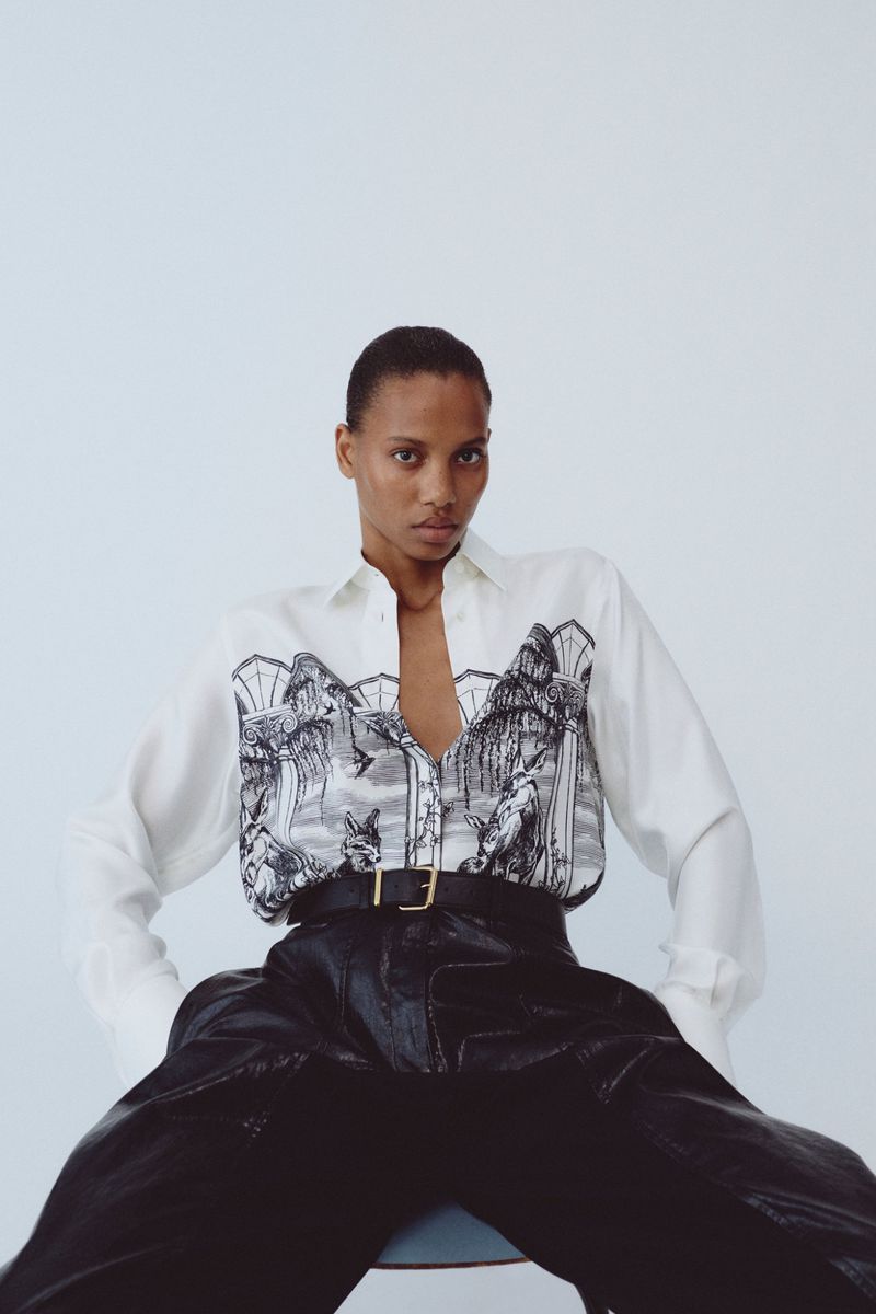 Model Cailane Oliveira seated in an illustrated white shirt and black leather trousers against a white studio background.