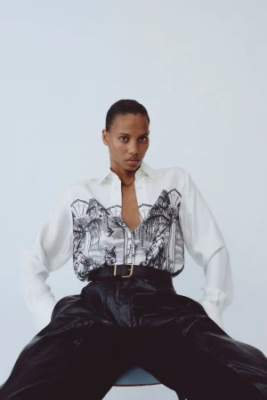 Model Cailane Oliveira seated in an illustrated white shirt and black leather trousers against a white studio background.