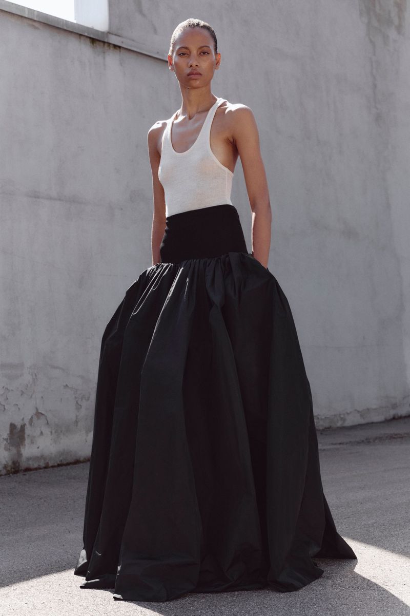 Model wearing a white scoop-neck tank top and a voluminous black floor-length skirt standing outdoors against a concrete wall.