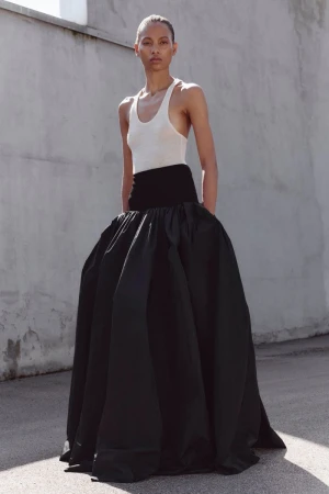 Model wearing a white scoop-neck tank top and a voluminous black floor-length skirt standing outdoors against a concrete wall.