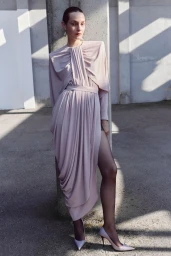 Model Clara Denison poses in a draped blush pink dress with a high slit against a concrete pillar in direct sunlight.