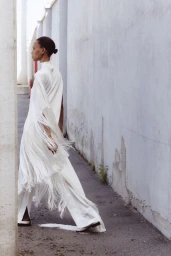 Model Cailane Oliveira walking outdoors in a flowing white fringe dress and wide-leg trousers for Stella McCartney Resort 2027.