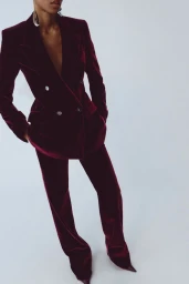 Model posing in a tailored burgundy velvet pantsuit and pointed heels against a white studio background.
