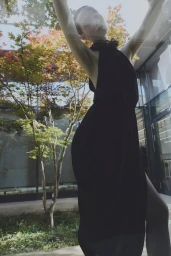 Model posing with arms raised wearing a sleeveless black halter dress in front of a tree and modern glass building.