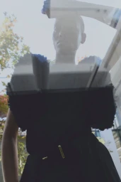 Low-angle shot of a model with short hair wearing a black belted garment seen through glass reflections of foliage and sky.