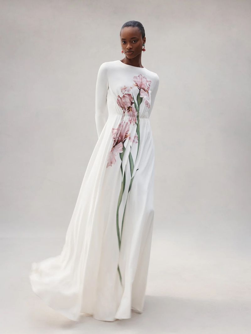 Model Mayowa Nicholas wearing a long-sleeve white floor-length gown with a large pink floral print against a neutral studio backdrop.