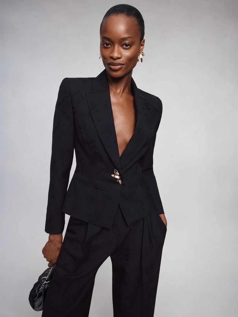 Model Mayowa Nicholas wearing a tailored black blazer with decorative brooch button and black pleated trousers against a grey backdrop.