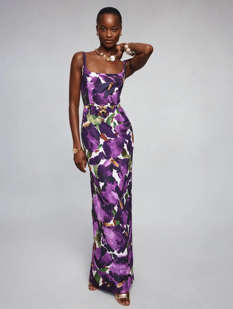 Model Mayowa Nicholas wears a purple floral maxi dress with gold jewelry against a grey studio backdrop for Oscar de la Renta.