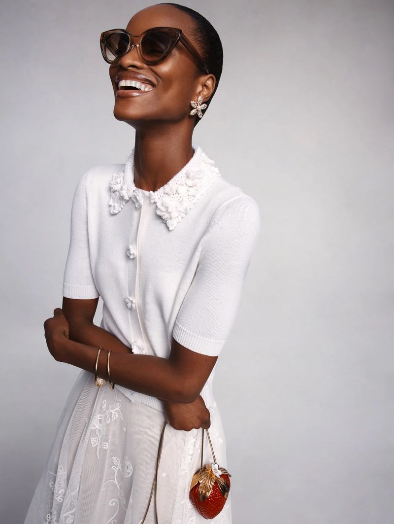 Mayowa Nicholas smiling in a white knit cardigan, cat-eye sunglasses, and holding a strawberry clutch on a studio background.