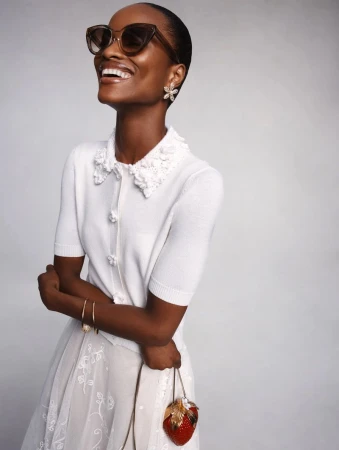 Mayowa Nicholas smiling in a white knit cardigan, cat-eye sunglasses, and holding a strawberry clutch on a studio background.