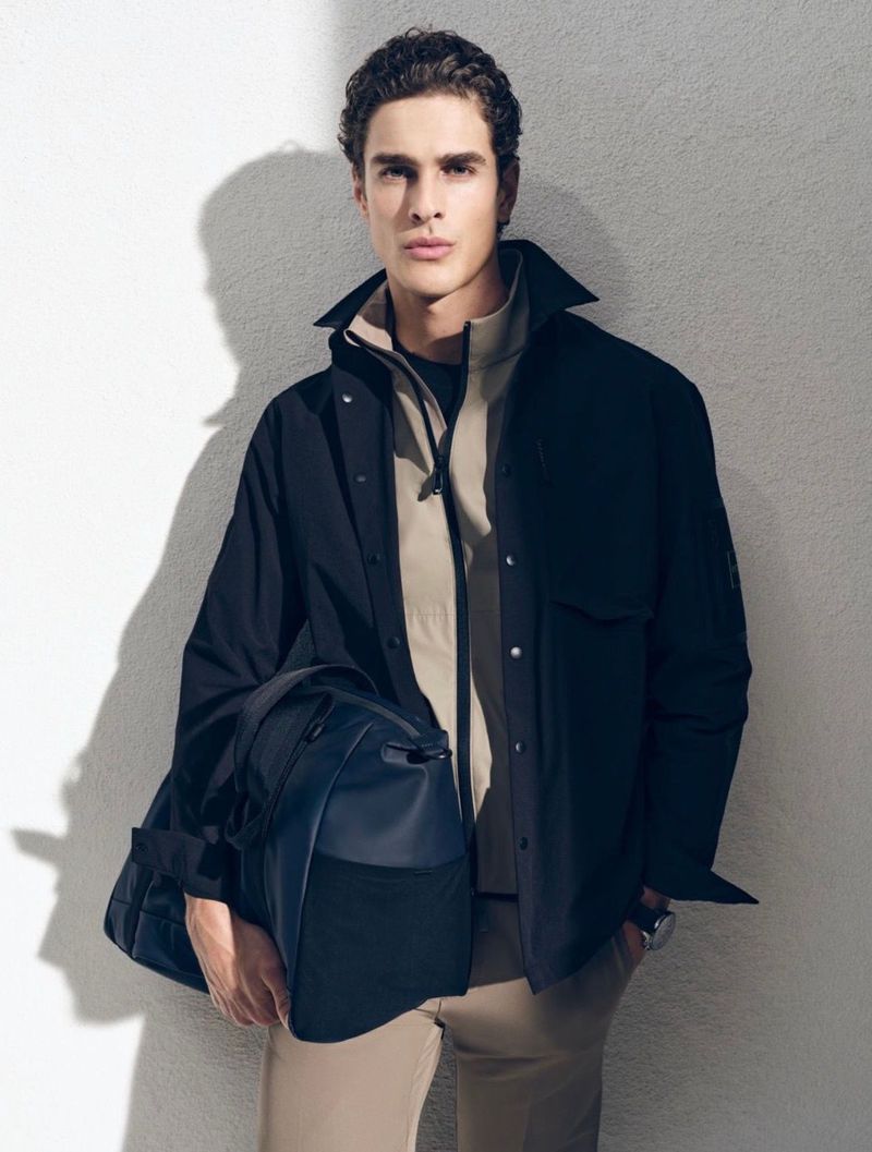 Model Max M holding a dark duffle bag while wearing an open black overshirt and beige zip jacket against a textured wall.