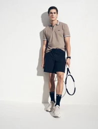 Model Max M in a taupe polo shirt and black athletic shorts holding a tennis racket against a white studio backdrop.