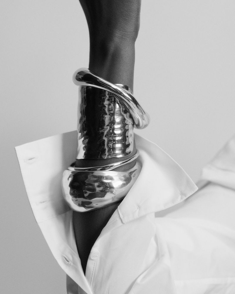 Close-up of a model's arm wearing sculptural silver cuff bracelets paired with an unbuttoned white collared shirt cuff.
