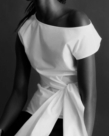 Model Maty Fall wears an off-the-shoulder white tied top against a dark studio background in black and white.