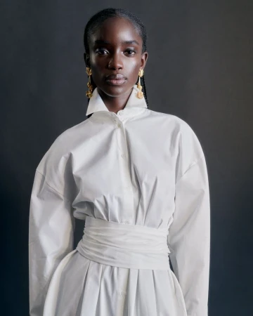Model Maty Fall posing in a crisp white collared dress with a gathered waistband and gold drop earrings against a dark backdrop.