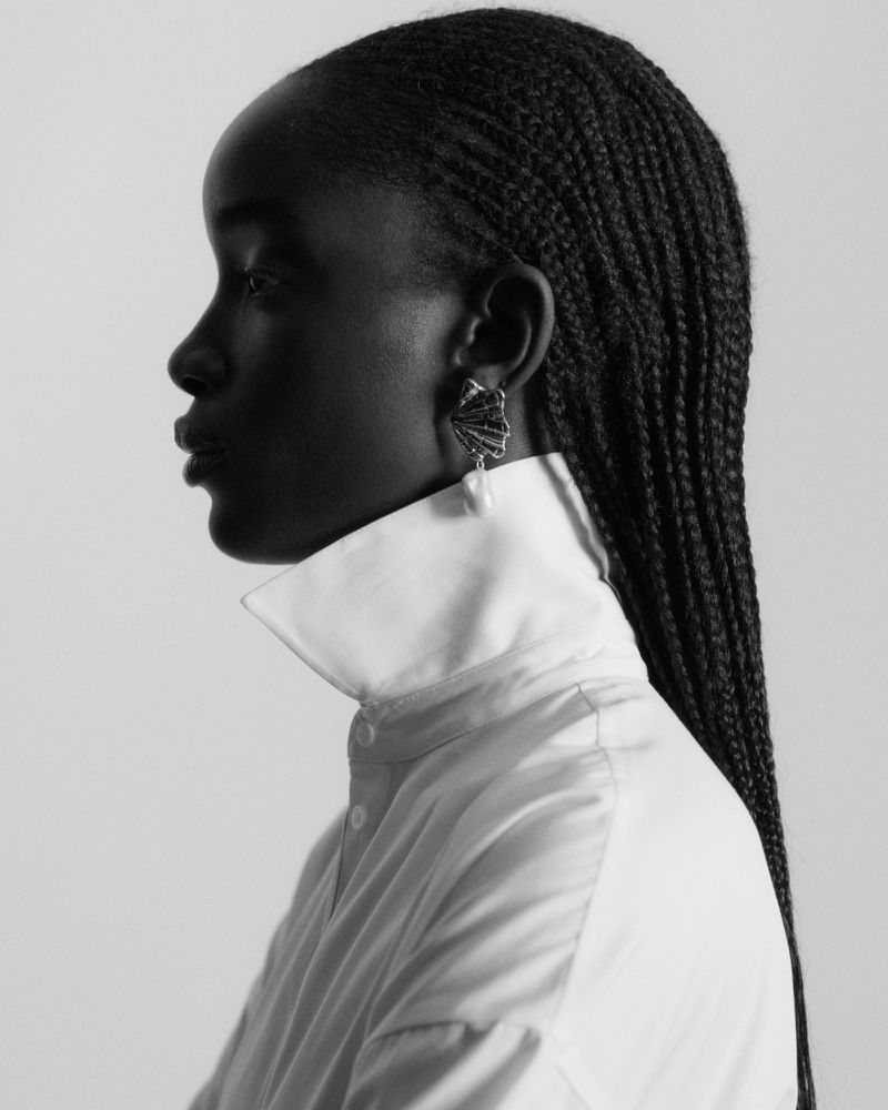 Maty Fall in profile wearing a high-collared white shirt and seashell pearl earring against a pale studio backdrop.