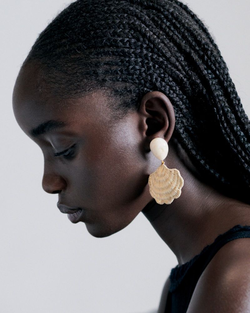 Model Maty Fall in profile wearing a cream shell drop earring and braided hair against a plain studio background.
