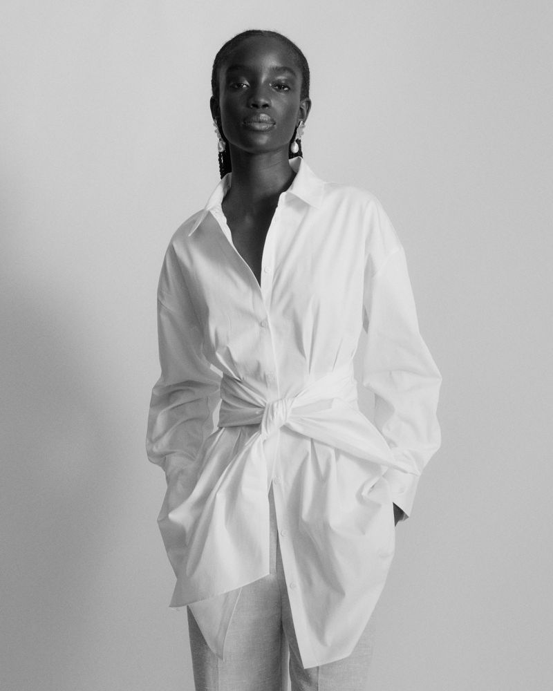 Model Maty Fall posing in a tailored white wrap shirt tied at the waist against a neutral studio backdrop in black and white.