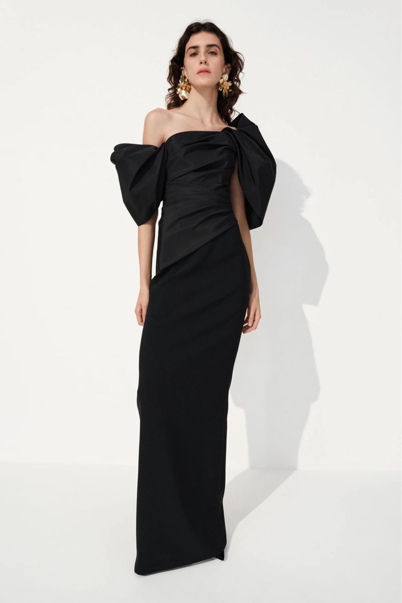 Model wearing a black off-the-shoulder evening gown with dramatic puffed sleeves and gold statement earrings against a white studio wall.