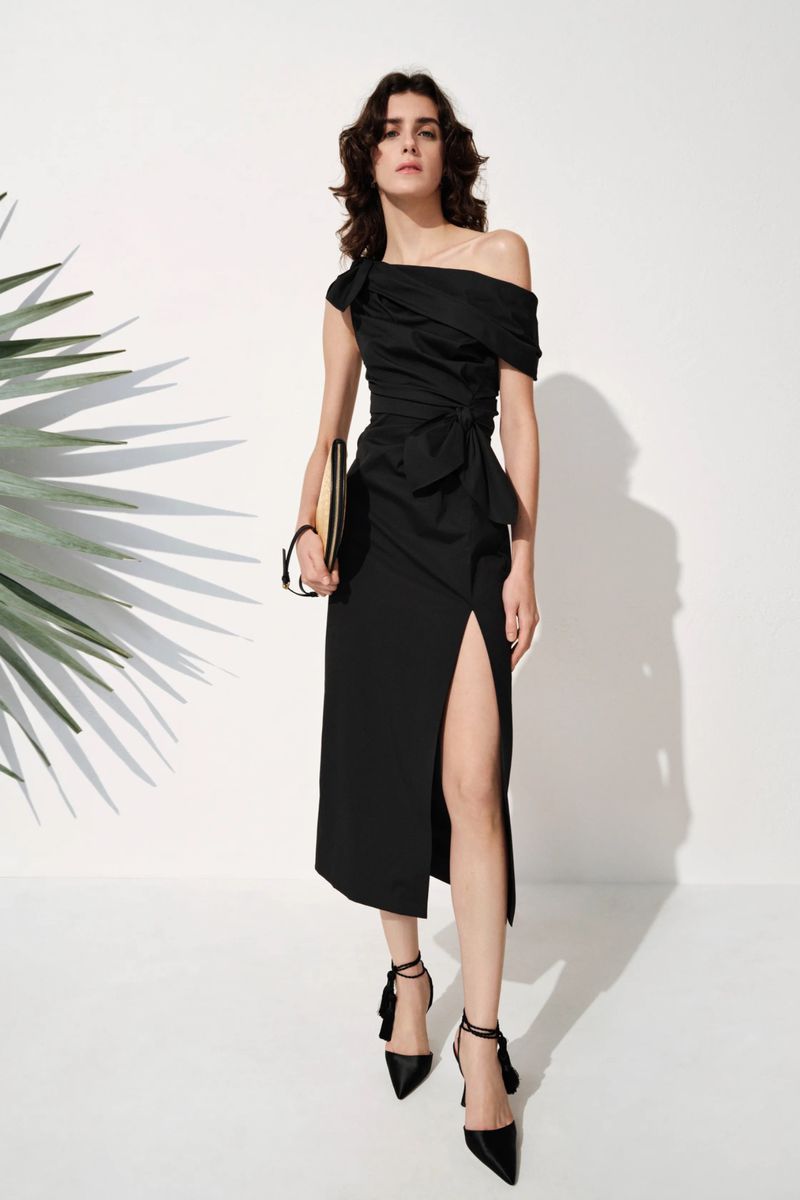 Model wearing a black off-the-shoulder slit dress and pointed heels against a white studio wall with palm shadows.