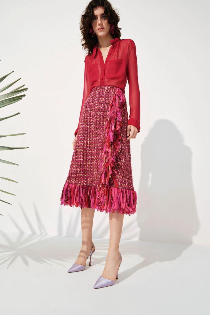 Model Julia Barucci wearing a sheer red blouse, fringed pink tweed skirt, and lilac heels against a white backdrop.