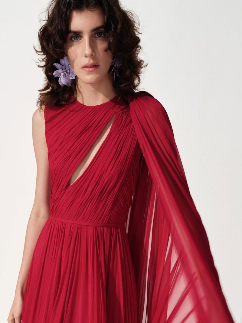 Model wearing a pleated red dress with a draped cape sleeve and large purple flower earrings against a plain white background