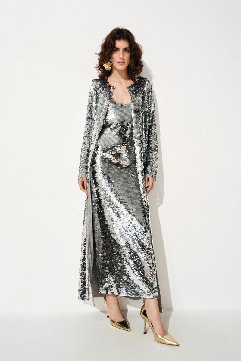 Model Julia Barucci in a silver sequined maxi dress and matching long coat with metallic gold heels against a white backdrop.