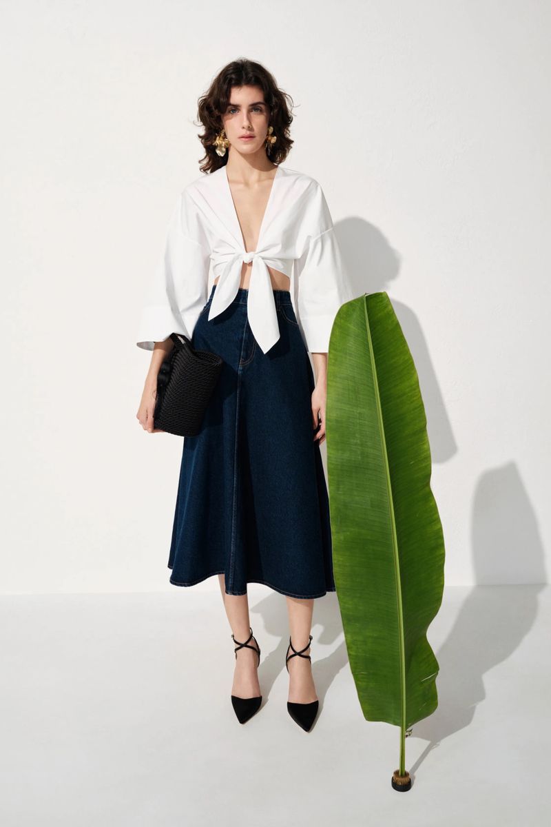 Model Julia Barucci in a white tie-front blouse and navy denim skirt beside a large green palm leaf against a white studio backdrop.