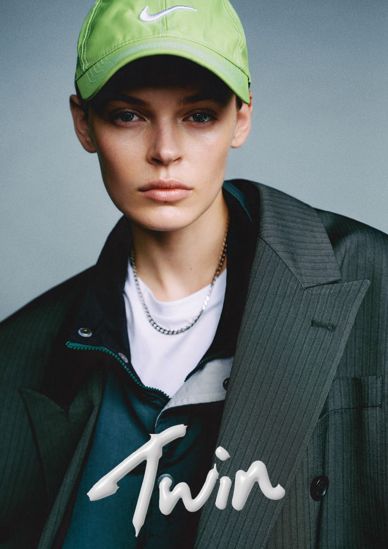 Model Cara Taylor wearing a bright lime green baseball cap and an oversized dark pinstripe blazer against a muted grey background.