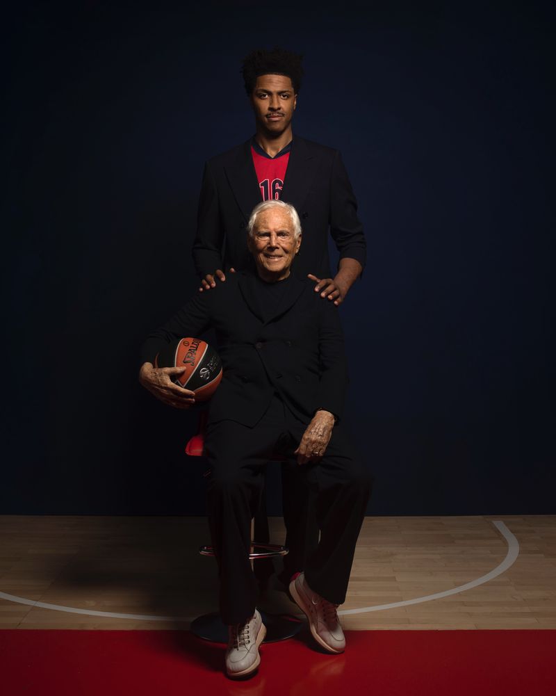 Giorgio Armani seated with a basketball, and standing next to a younger basketball player in a portrait, in front of a dark background.