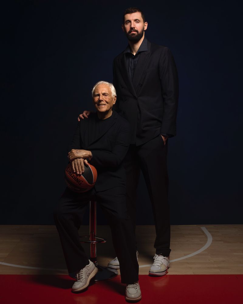 Portrait of a smiling senior adult male sitting and holding a basketball, standing next to a tall adult male in a dark suit.