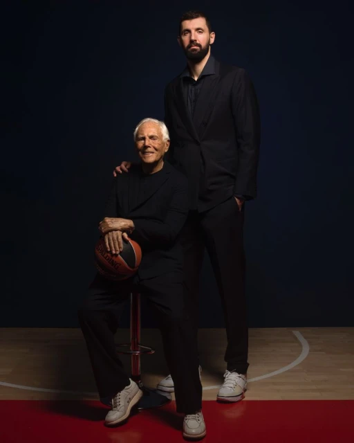 Portrait of a smiling senior adult male sitting and holding a basketball, standing next to a tall adult male in a dark suit.
