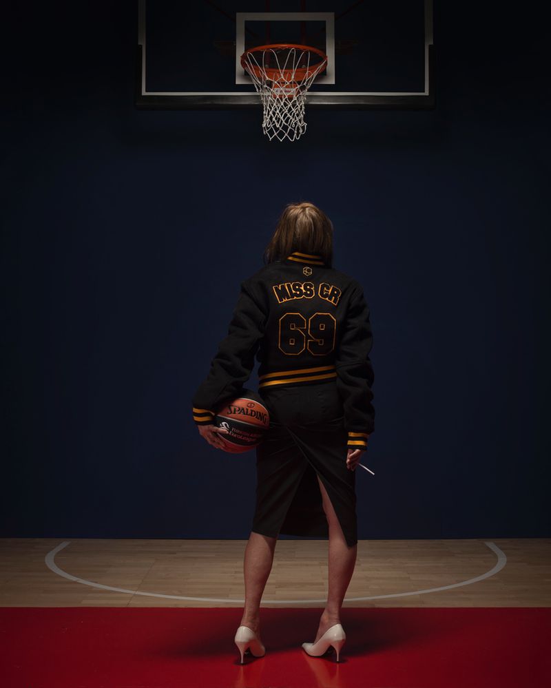 Woman in varsity jacket holds a basketball on a court with hoop, back to camera, against a dark blue background in an indoor setting.