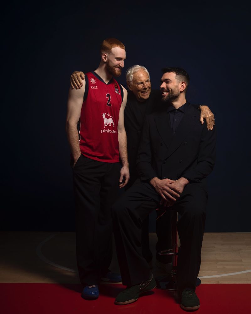 Three men are shown indoors, the man on the left in a red basketball jersey, the man in the center with white hair, and the man on the right in a black suit.