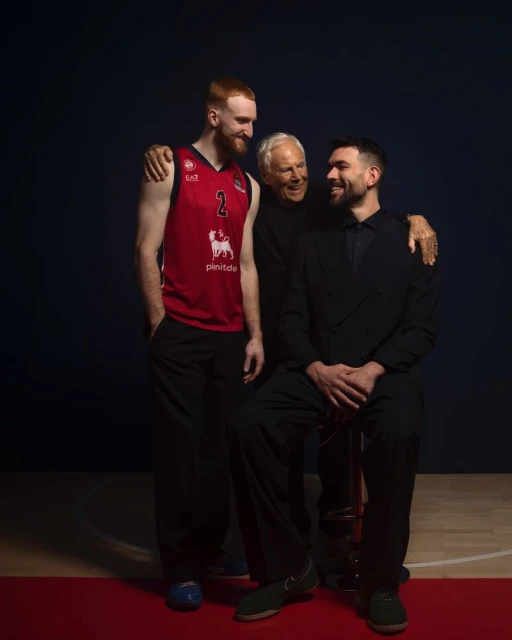 Three men are shown indoors, the man on the left in a red basketball jersey, the man in the center with white hair, and the man on the right in a black suit.