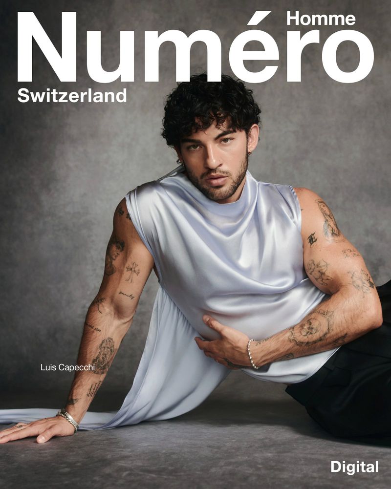 Luis Capecchi wearing a draped sleeveless lavender satin top and black trousers against a textured grey studio backdrop.