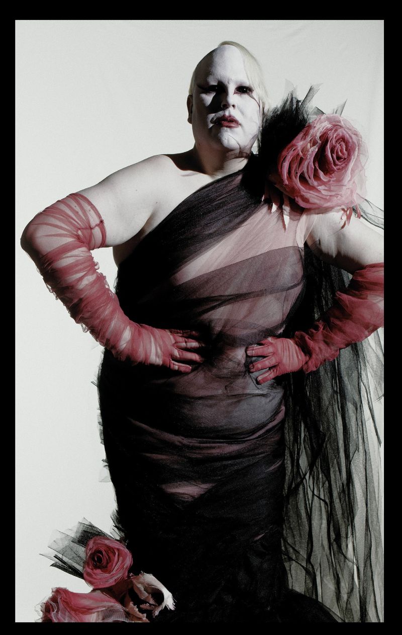 Avant-garde figure wearing a sheer black tulle gown with pink floral corsages and red mesh opera gloves against a white studio backdrop.