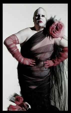 Avant-garde figure wearing a sheer black tulle gown with pink floral corsages and red mesh opera gloves against a white studio backdrop.