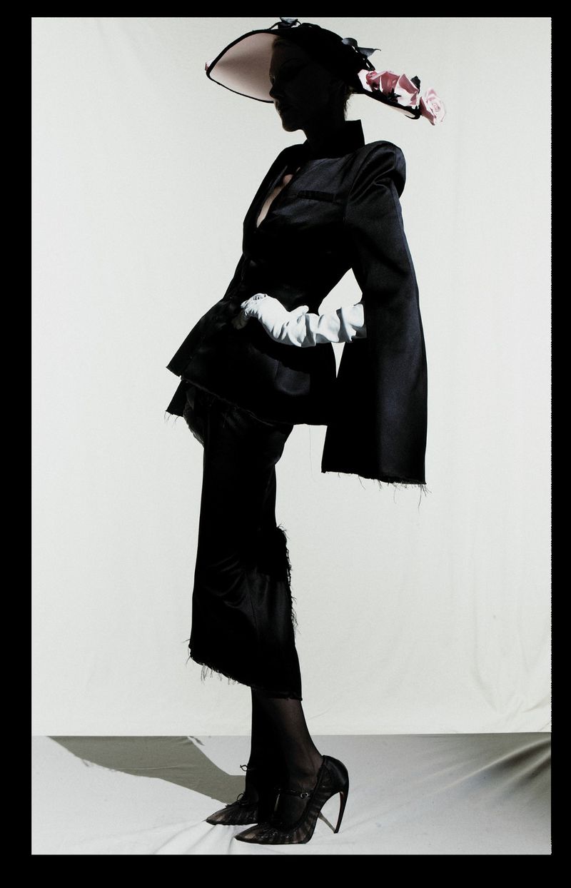 Model posing in a sculptural black suit, white gloves, floral-accented hat, and high heels against a white studio background.