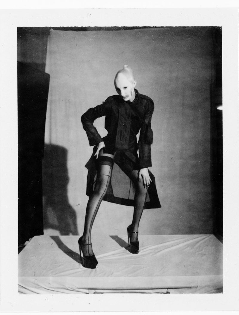 Black and white portrait of a model in a sheer dark coat, seamed stockings, and platform heels posing on a low platform in a studio.