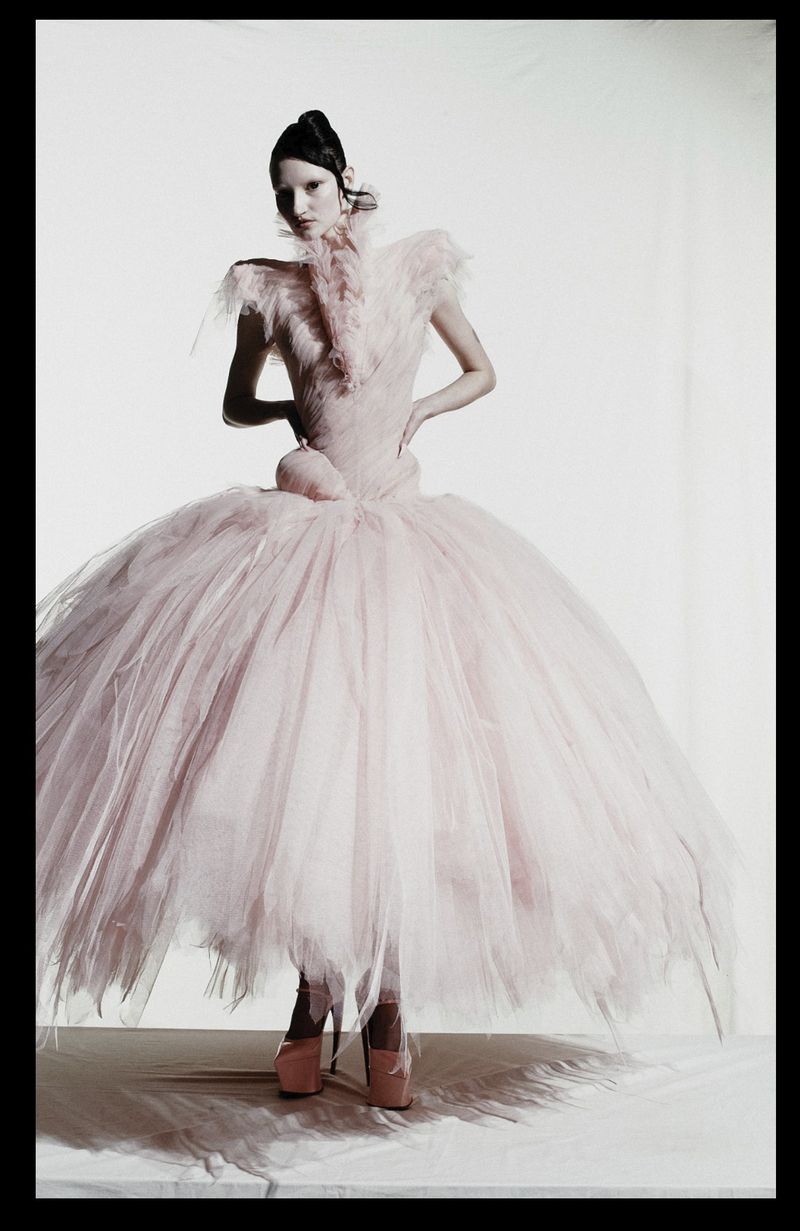 Model wearing an expansive blush pink tulle gown and platform heels posing against a white studio backdrop.